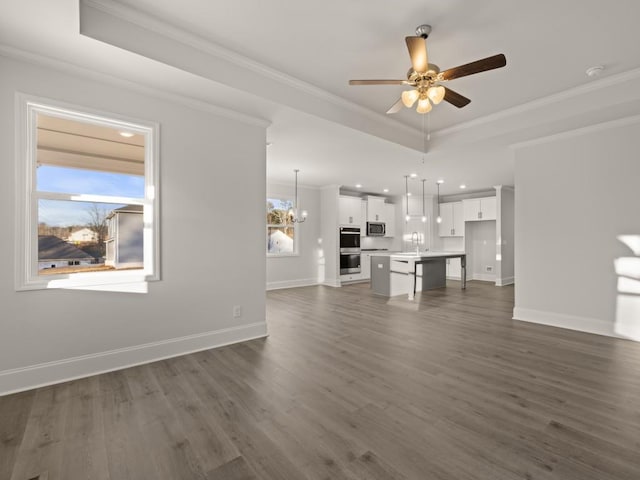 unfurnished living room with sink, ornamental molding, dark hardwood / wood-style floors, a tray ceiling, and ceiling fan with notable chandelier