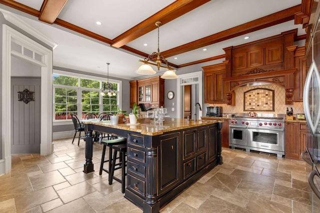 kitchen with light stone countertops, hanging light fixtures, crown molding, range with two ovens, and a kitchen island with sink