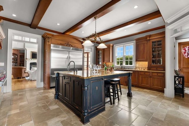 kitchen featuring light stone counters, stainless steel built in refrigerator, a kitchen island with sink, beamed ceiling, and hanging light fixtures