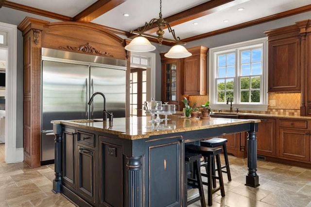 kitchen with light stone countertops, tasteful backsplash, stainless steel built in fridge, hanging light fixtures, and an island with sink