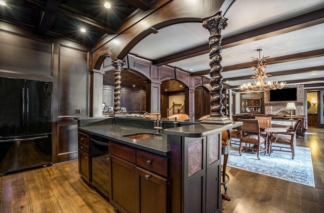 kitchen with sink, beamed ceiling, decorative columns, an island with sink, and black appliances