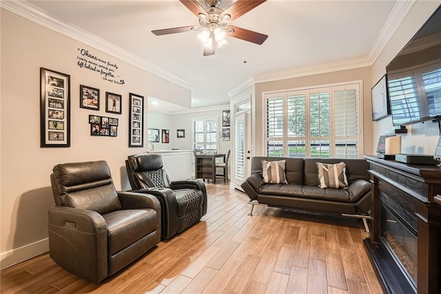 living area featuring ceiling fan, a glass covered fireplace, baseboards, light wood-type flooring, and ornamental molding