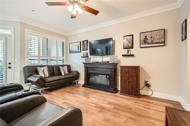living room with crown molding, a glass covered fireplace, light wood-style flooring, a ceiling fan, and baseboards