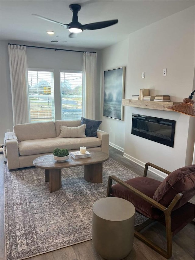 living area featuring a ceiling fan, a glass covered fireplace, baseboards, and wood finished floors