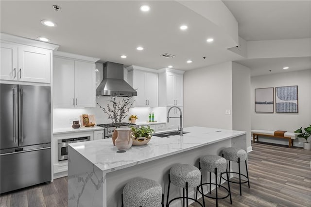 kitchen featuring stainless steel appliances, a sink, white cabinets, wall chimney range hood, and a center island with sink