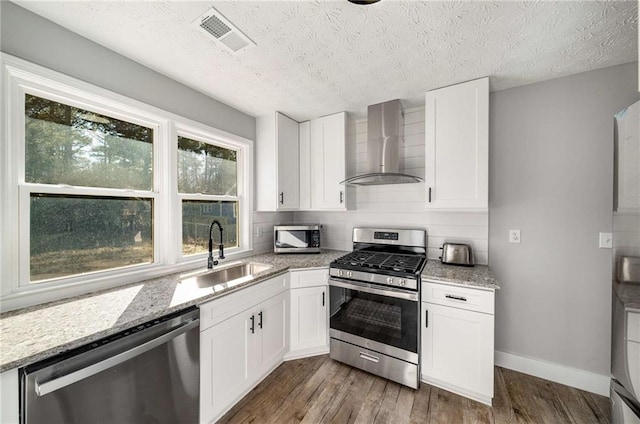 kitchen with light stone countertops, appliances with stainless steel finishes, white cabinets, wall chimney range hood, and sink