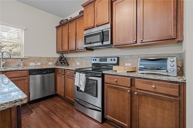 kitchen with appliances with stainless steel finishes, a sink, dark wood-style flooring, brown cabinetry, and light stone counters