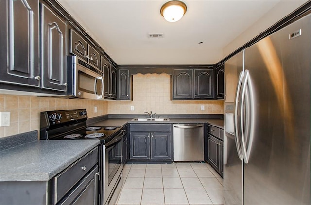 kitchen with appliances with stainless steel finishes, a sink, light tile patterned flooring, dark countertops, and decorative backsplash