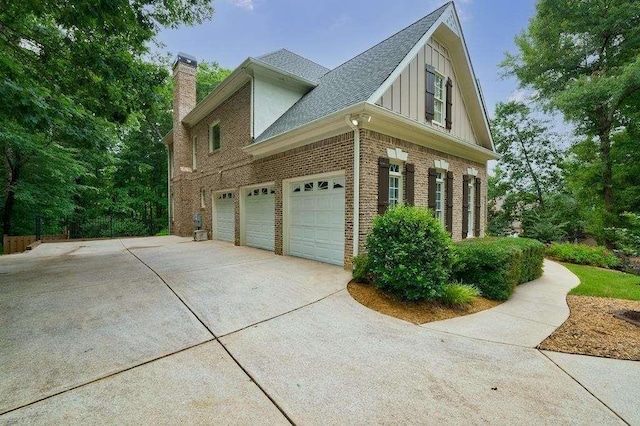 view of home's exterior with driveway, roof with shingles, an attached garage, brick siding, and a chimney