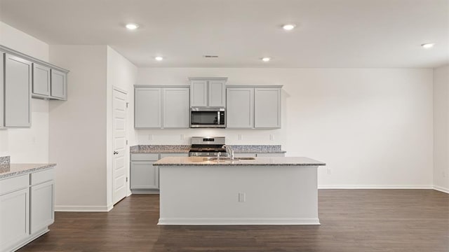 kitchen with dark wood finished floors, appliances with stainless steel finishes, light stone counters, a kitchen island with sink, and gray cabinets