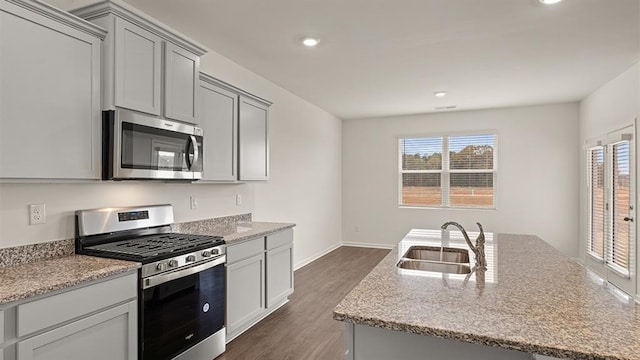 kitchen with light stone counters, a center island with sink, appliances with stainless steel finishes, dark wood-type flooring, and a sink