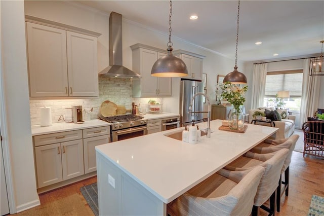 kitchen featuring stainless steel appliances, wall chimney exhaust hood, a sink, a center island with sink, and backsplash