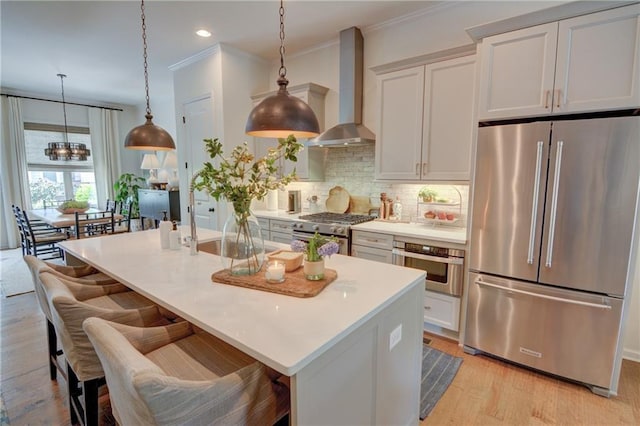 kitchen with high end appliances, wall chimney exhaust hood, light wood finished floors, backsplash, and an island with sink