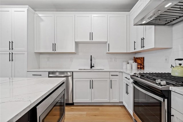 kitchen with light wood-type flooring, a sink, stainless steel appliances, wall chimney exhaust hood, and white cabinets