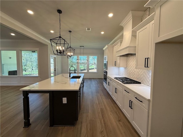 kitchen with tasteful backsplash, custom exhaust hood, stainless steel gas cooktop, hanging light fixtures, and an island with sink