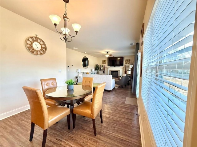 dining space with baseboards, wood finished floors, a glass covered fireplace, and ceiling fan with notable chandelier