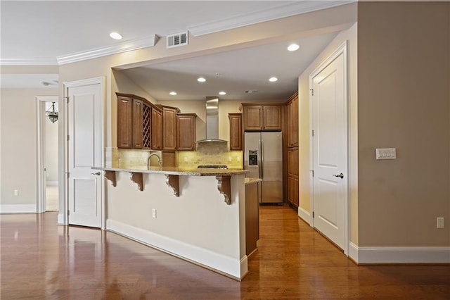 kitchen with stainless steel fridge with ice dispenser, wall chimney range hood, a peninsula, a breakfast bar, and light stone counters