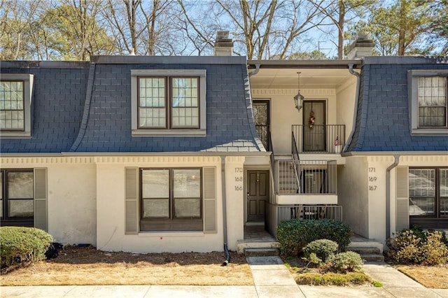 multi unit property with a shingled roof, a chimney, mansard roof, and stucco siding