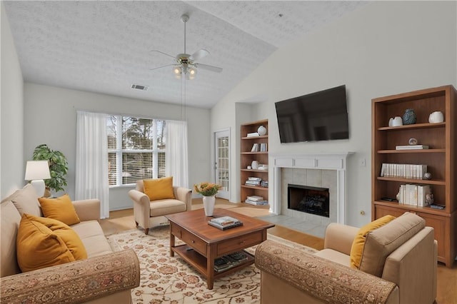 living room featuring lofted ceiling, a tile fireplace, ceiling fan, a textured ceiling, and built in shelves