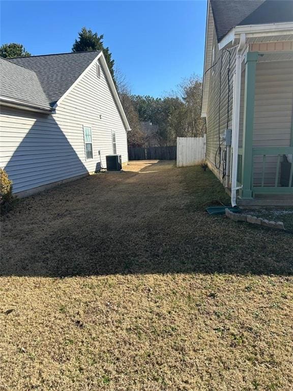 view of side of home with a lawn, central AC, roof with shingles, and fence