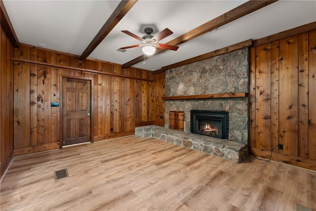 unfurnished living room with a stone fireplace, wooden walls, light hardwood / wood-style floors, ceiling fan, and beam ceiling