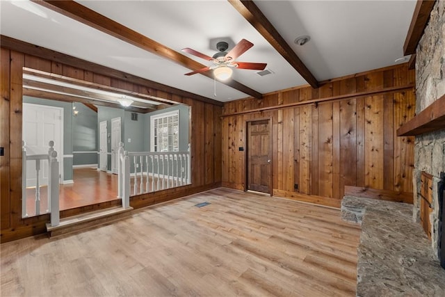 unfurnished room featuring ceiling fan, beam ceiling, wooden walls, light hardwood / wood-style flooring, and a stone fireplace