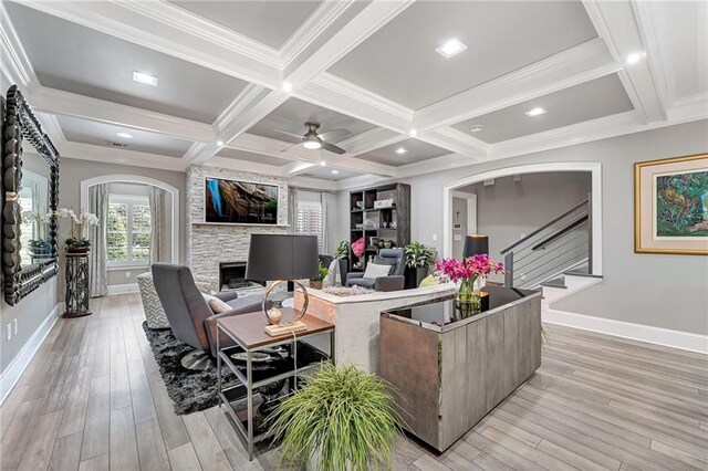 living room featuring ceiling fan, coffered ceiling, a stone fireplace, light hardwood / wood-style flooring, and beamed ceiling