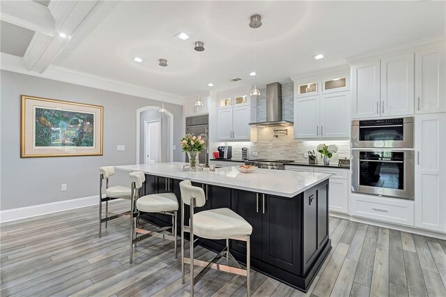 kitchen with white cabinets, wall chimney exhaust hood, light wood-type flooring, an island with sink, and appliances with stainless steel finishes