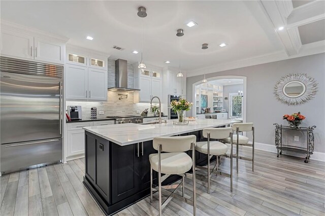 kitchen featuring white cabinetry, wall chimney exhaust hood, stainless steel appliances, an island with sink, and pendant lighting