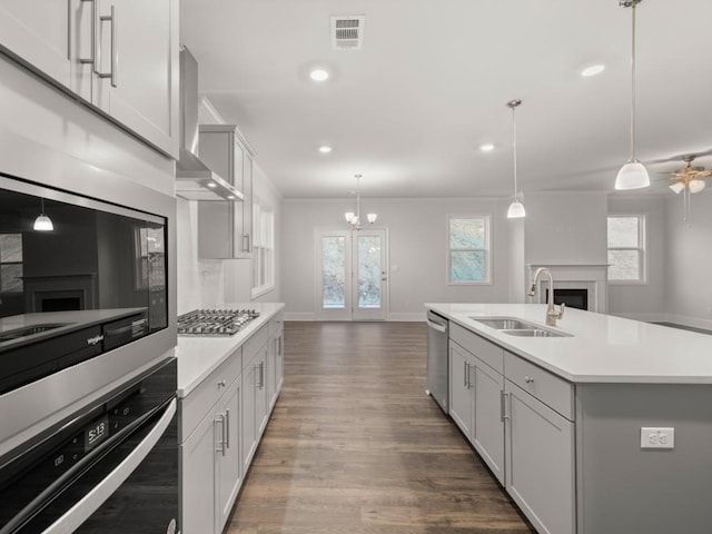 kitchen with decorative light fixtures, a center island with sink, sink, appliances with stainless steel finishes, and wall chimney exhaust hood