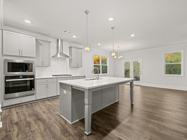 kitchen featuring sink, hanging light fixtures, appliances with stainless steel finishes, an island with sink, and wall chimney exhaust hood