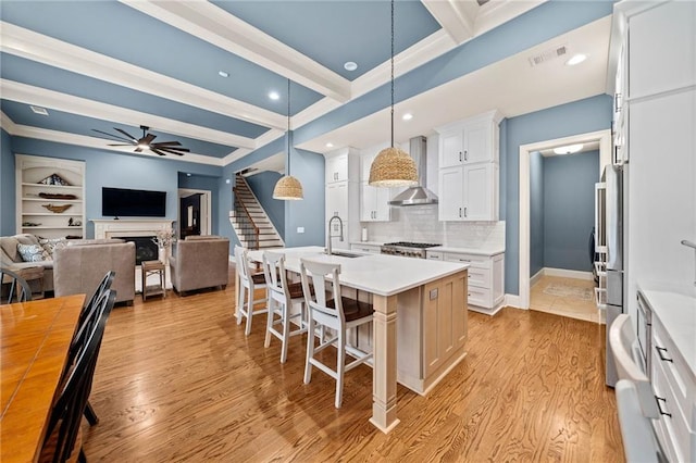 kitchen with white cabinets, a kitchen breakfast bar, hanging light fixtures, a center island with sink, and wall chimney range hood