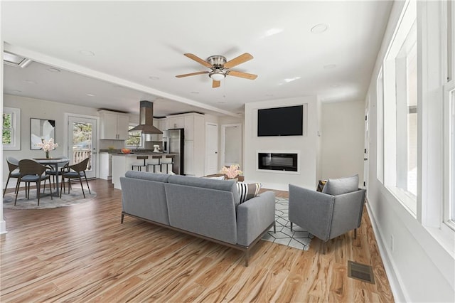 living area featuring a ceiling fan, light wood-type flooring, visible vents, and a glass covered fireplace