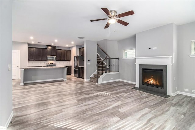 unfurnished living room featuring light wood-style floors, a fireplace with flush hearth, baseboards, ceiling fan, and stairs