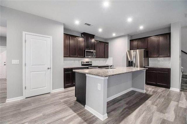 kitchen featuring appliances with stainless steel finishes, backsplash, light wood-style floors, a center island with sink, and dark brown cabinets