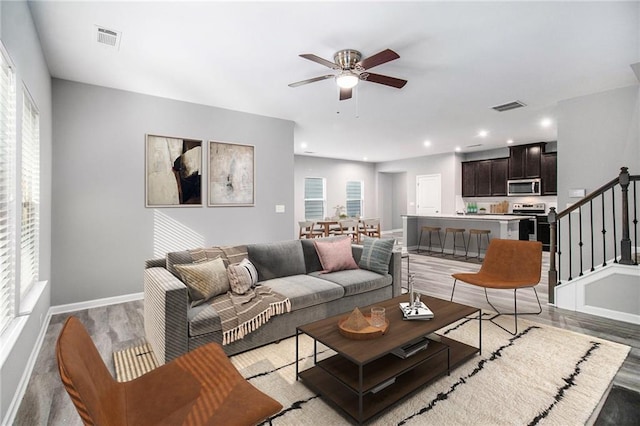 living area with light wood-style floors, a ceiling fan, stairs, baseboards, and plenty of natural light