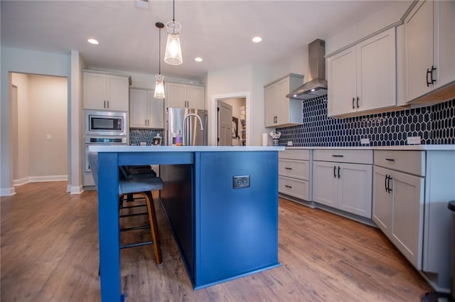 kitchen featuring hanging light fixtures, wall chimney range hood, stainless steel appliances, a center island with sink, and light wood-type flooring