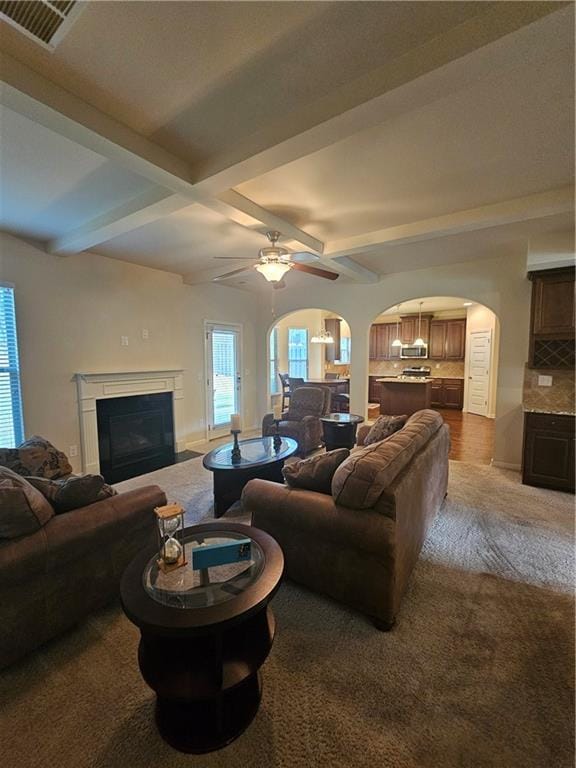 living area with carpet floors, arched walkways, beam ceiling, ceiling fan, and a glass covered fireplace