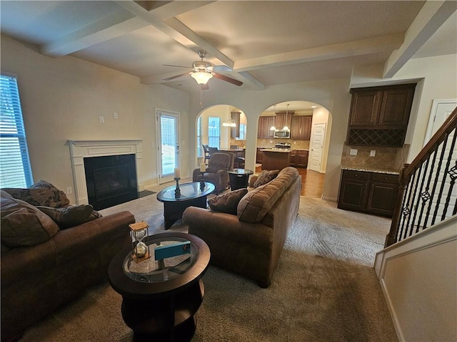 living area featuring arched walkways, beamed ceiling, light colored carpet, a ceiling fan, and a fireplace with flush hearth