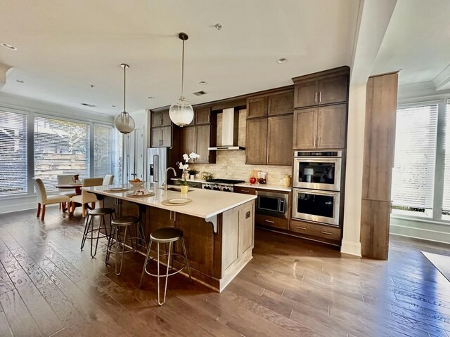 kitchen with dark wood-type flooring, stainless steel appliances, wall chimney range hood, decorative light fixtures, and a kitchen island with sink