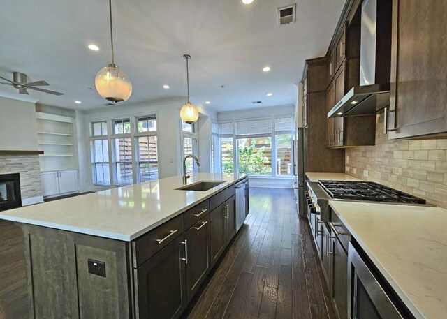 kitchen with wall chimney range hood, sink, hanging light fixtures, an island with sink, and a wealth of natural light