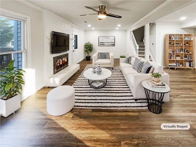 living room featuring ceiling fan, wood finished floors, stairs, a brick fireplace, and crown molding