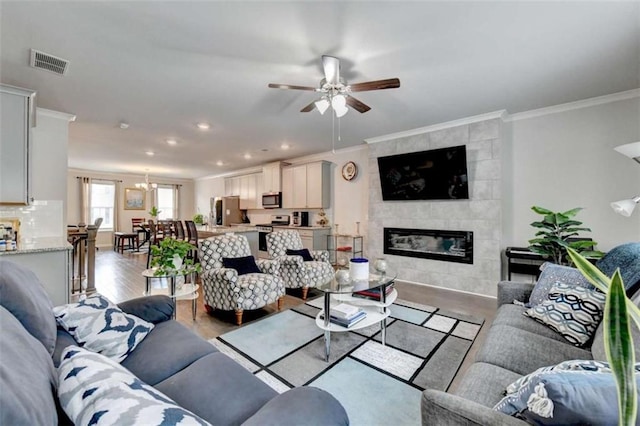 living room featuring visible vents, a tile fireplace, wood finished floors, crown molding, and ceiling fan with notable chandelier