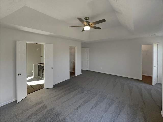 unfurnished bedroom featuring ensuite bathroom, a spacious closet, a raised ceiling, ceiling fan, and dark colored carpet