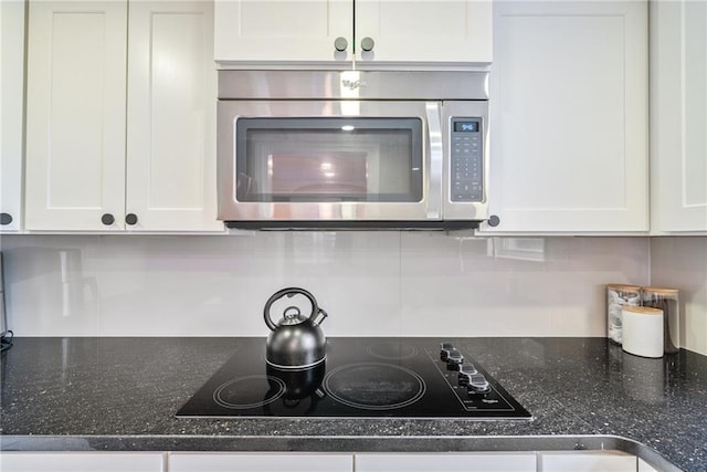 kitchen featuring white cabinetry, stainless steel microwave, black electric stovetop, and dark stone countertops