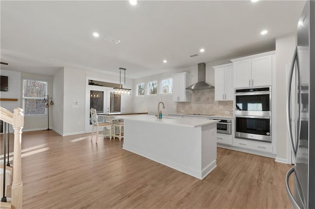 kitchen featuring appliances with stainless steel finishes, an island with sink, pendant lighting, wall chimney range hood, and white cabinets