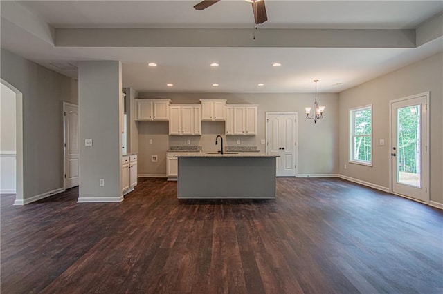 kitchen featuring an island with sink, ceiling fan with notable chandelier, dark wood-style floors, white cabinets, and baseboards