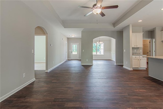 unfurnished living room featuring ceiling fan, baseboards, a tray ceiling, arched walkways, and dark wood-style flooring