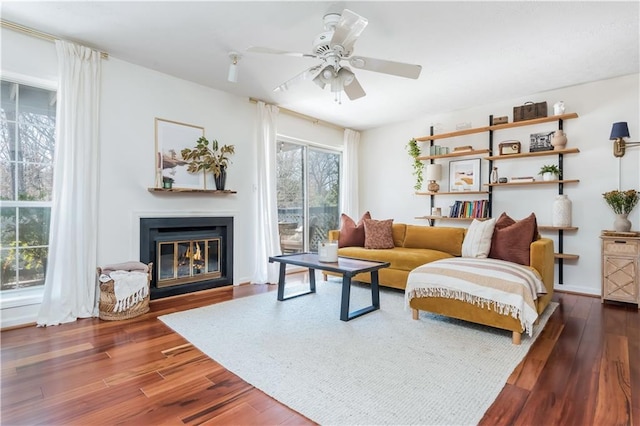 living area with a glass covered fireplace, dark wood finished floors, and a ceiling fan