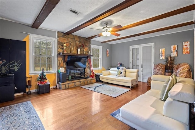 living room with a stone fireplace, hardwood / wood-style flooring, ceiling fan, a textured ceiling, and beamed ceiling